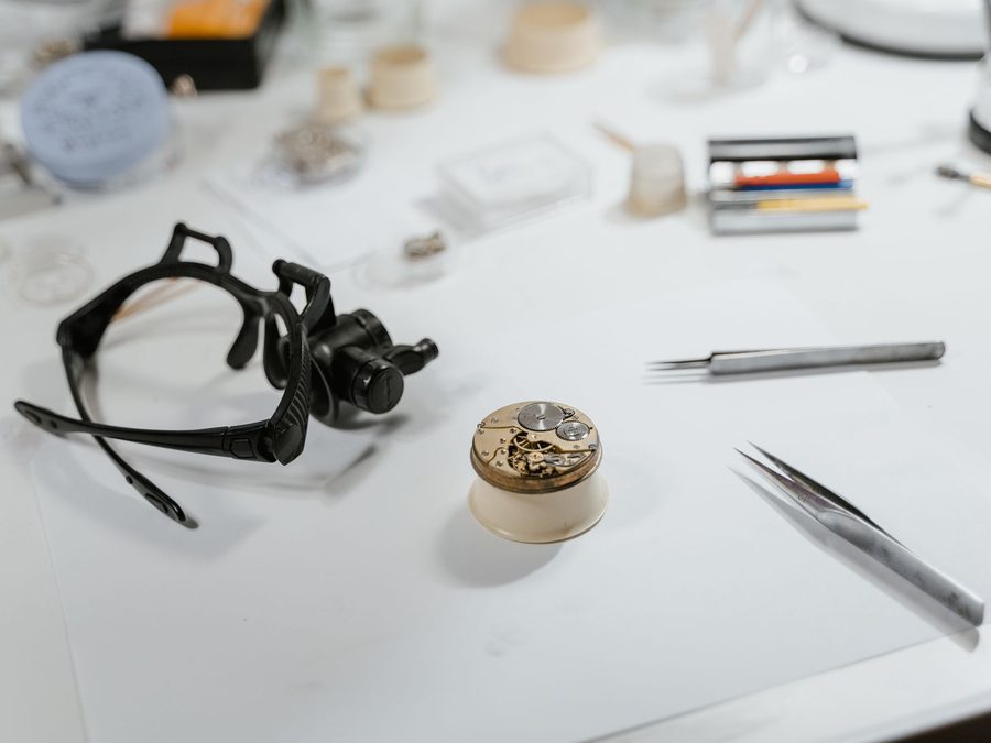 A watchmaker's white bench: a movement on its holder, a head loupe and two pairs of tweezers.