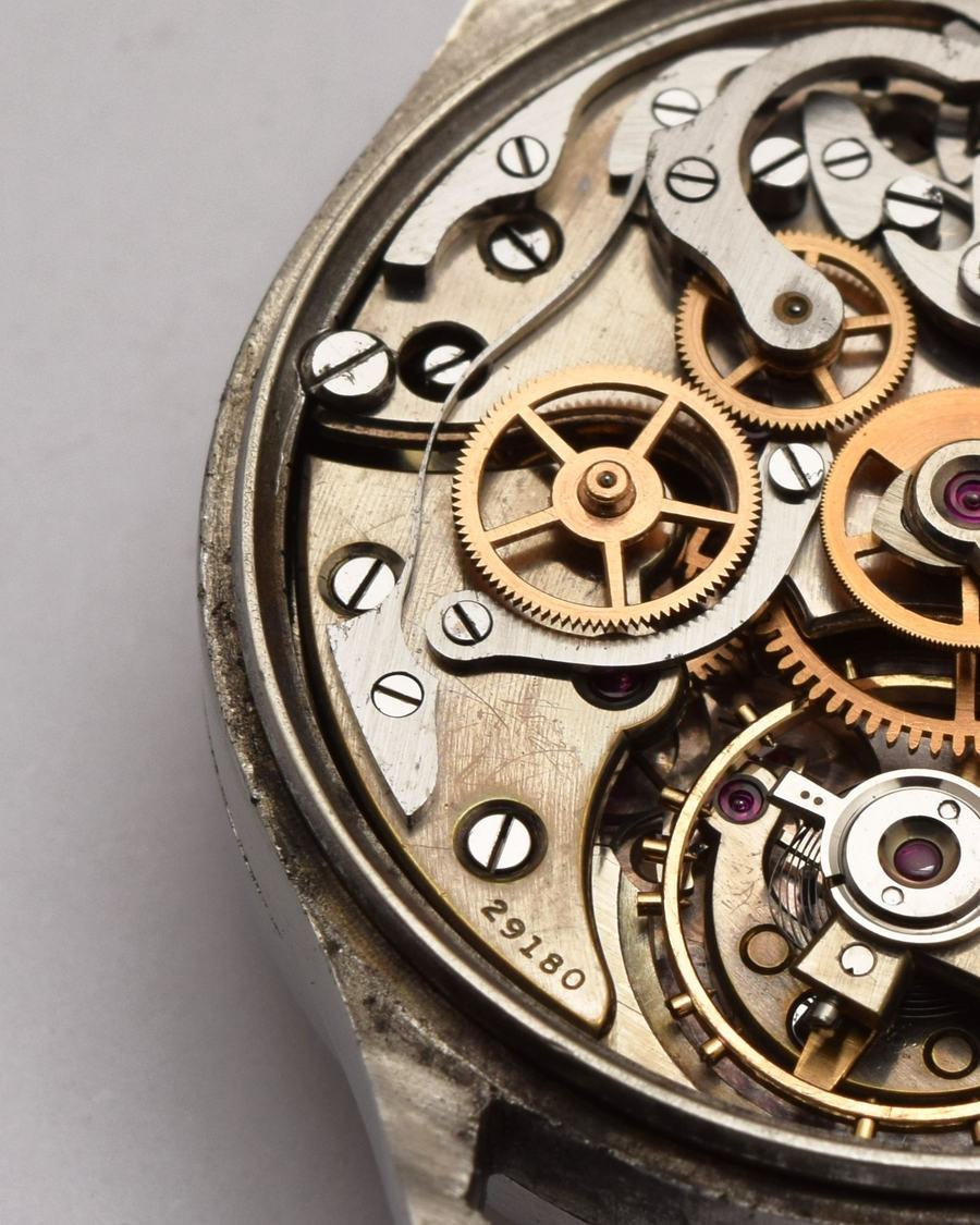 Close-up of a vintage chronograph movement: brass wheels, steel levers and ruby jewels inside a worn steel case.