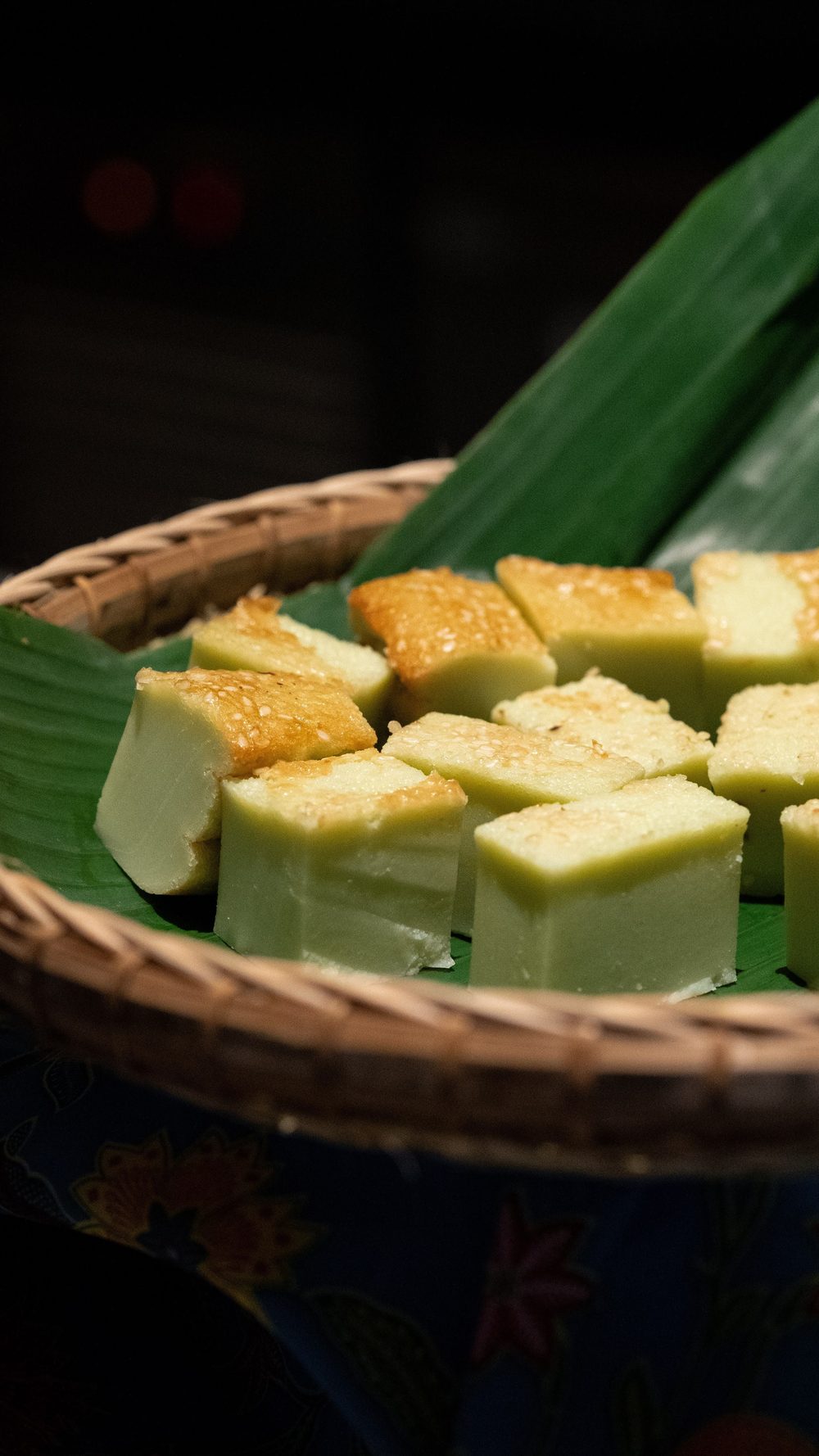 Squares of green kueh with toasted, sesame-flecked tops on banana leaf in a rattan basket, lit low against a dark room.