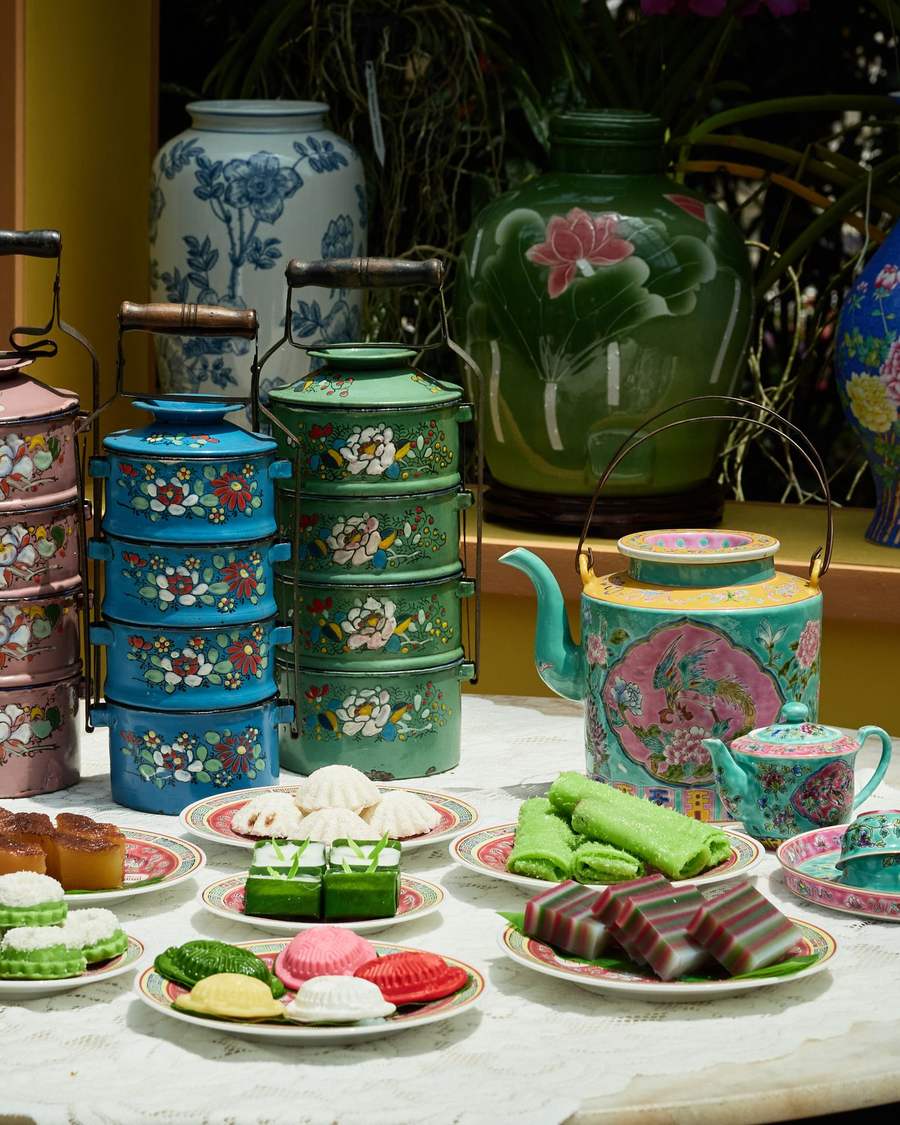 A marble table set with plates of colourful kueh, stacked enamel tingkat carriers in pink, blue and green, and a turquoise porcelain teapot painted with a phoenix.
