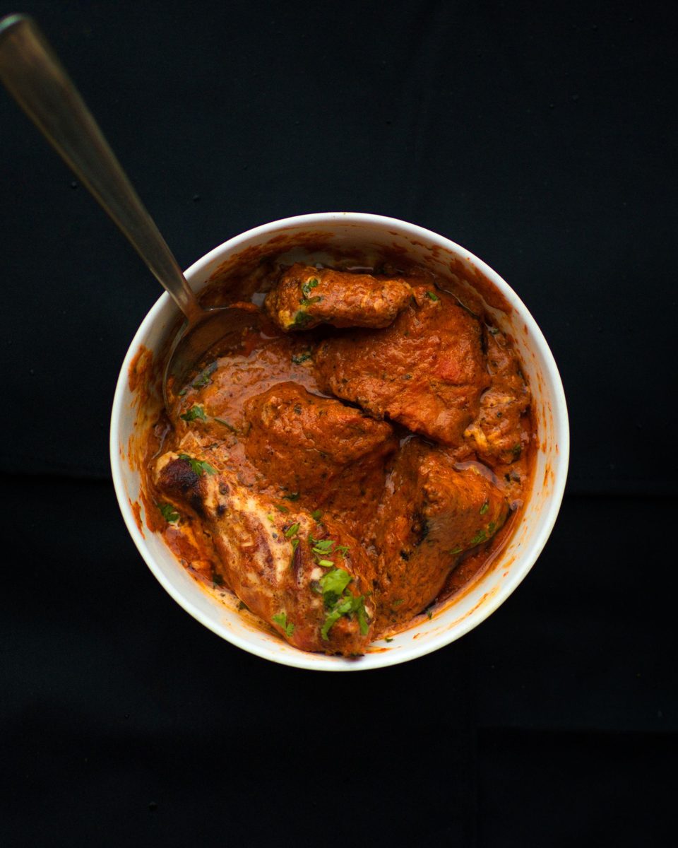 Looking down on a bowl of deep red chicken curry with a spoon resting in it, on a black background.