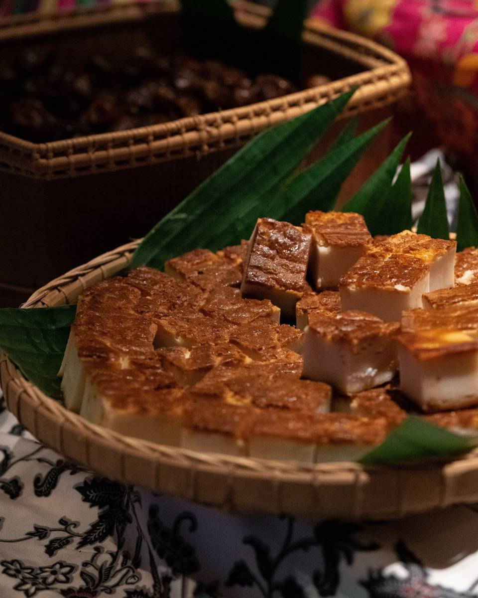 A rattan tray of golden baked kueh squares on banana leaves, set on a batik cloth.