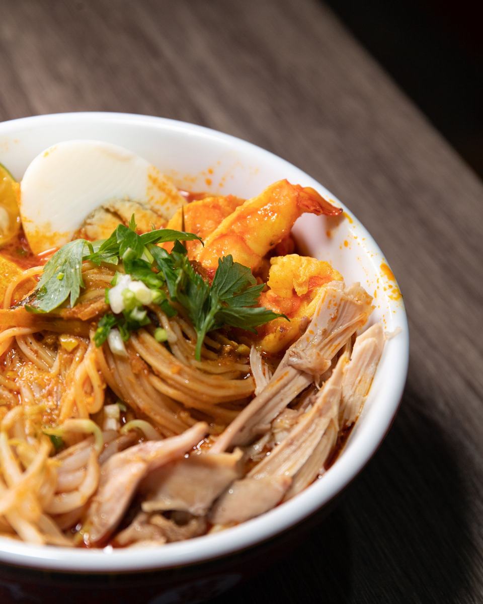 A white bowl of laksa: noodles in orange coconut gravy with prawns, half an egg, shredded chicken and fresh herbs.