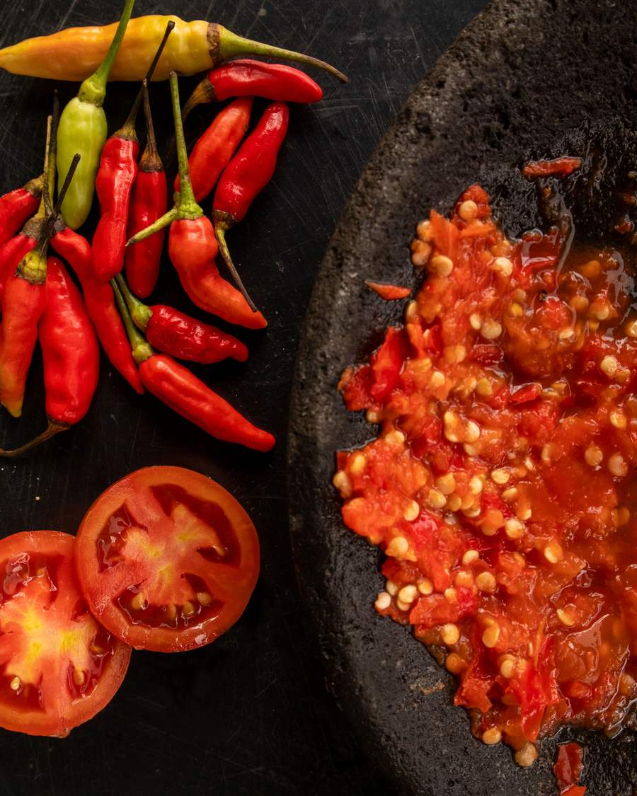 Red chillies and halved tomatoes beside a stone mortar of freshly pounded red sambal.
