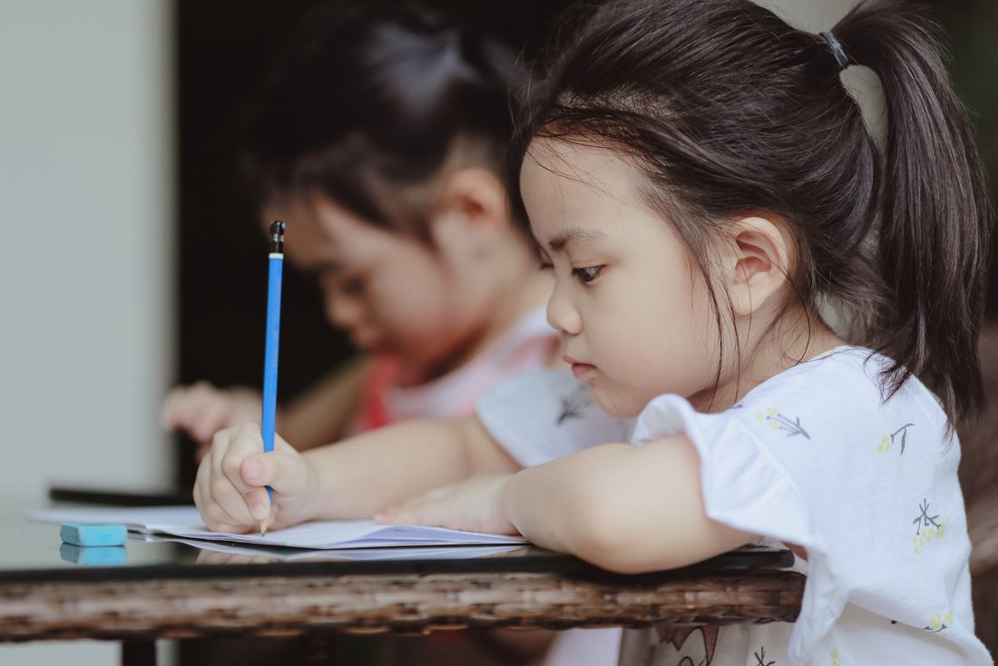 A young girl concentrating as she writes in her exercise book, with another child working beside her