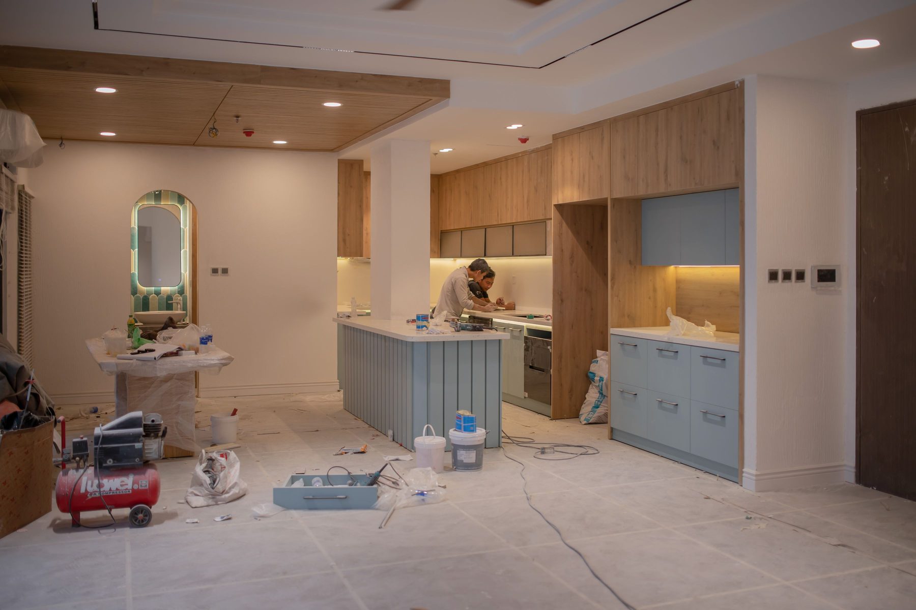 An open-plan kitchen mid-renovation: two workers fitting the worktop, new wood-grain and pale blue cabinets in place, tools and paint tins on the protected floor.
