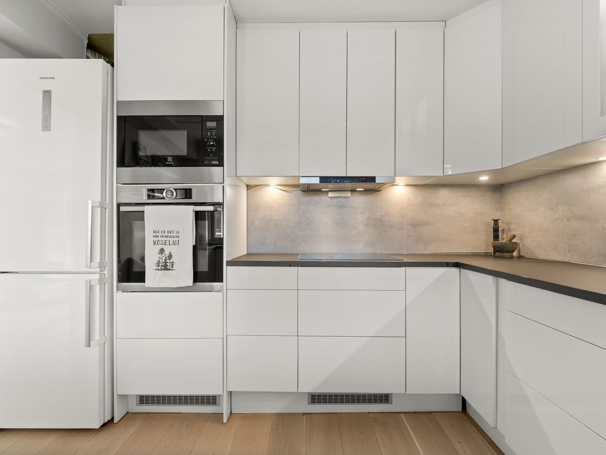 After: a white L-shaped kitchen with built-in oven, under-cabinet lights and light wood flooring.