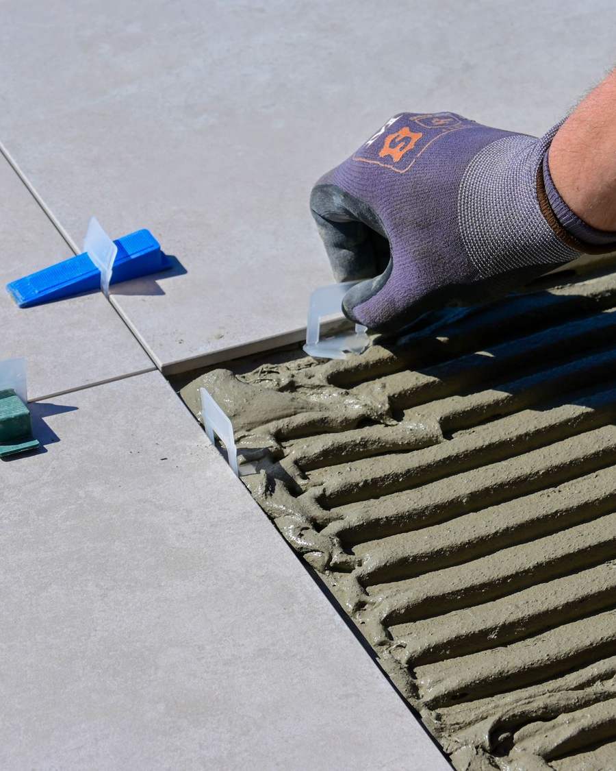 A gloved hand setting large grey floor tiles on combed adhesive, with blue levelling clips holding the joints flat.