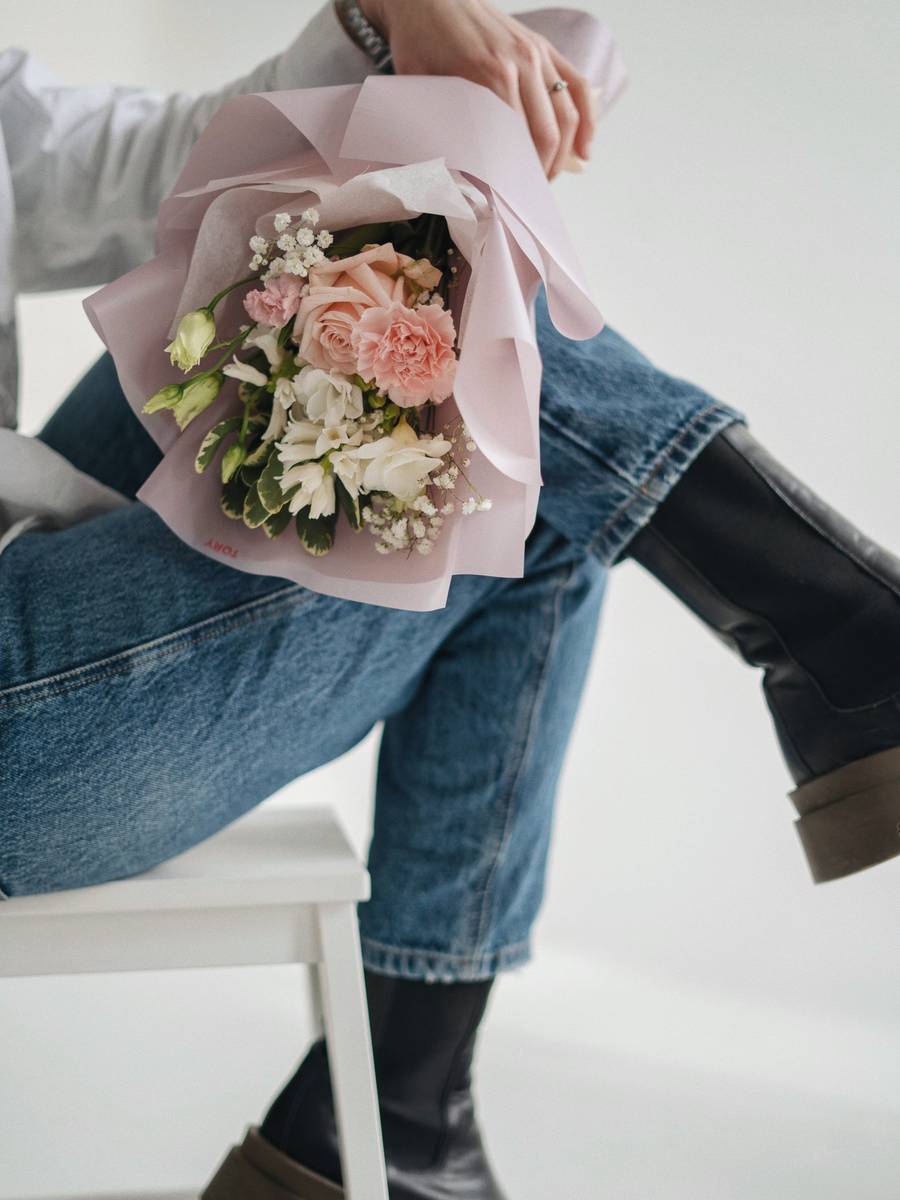 A hand-tied bouquet of peach roses and white spray flowers wrapped in pale pink paper, resting on someone's lap.