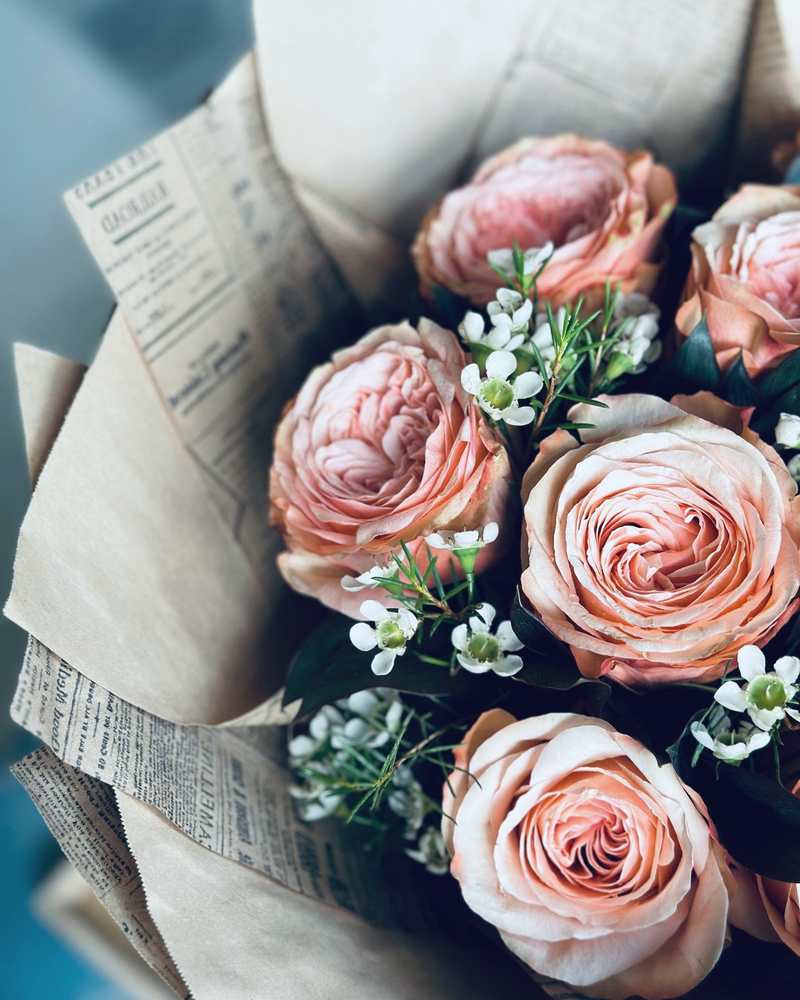 Peach garden roses and white waxflower wrapped in kraft and vintage newsprint, seen from above.