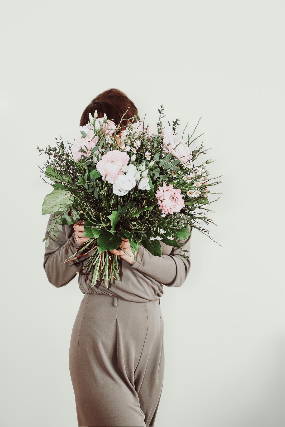 Someone holding a large, loose bouquet of pale pink ranunculus, white lisianthus, chamomile and trailing greenery, the flowers hiding their face.