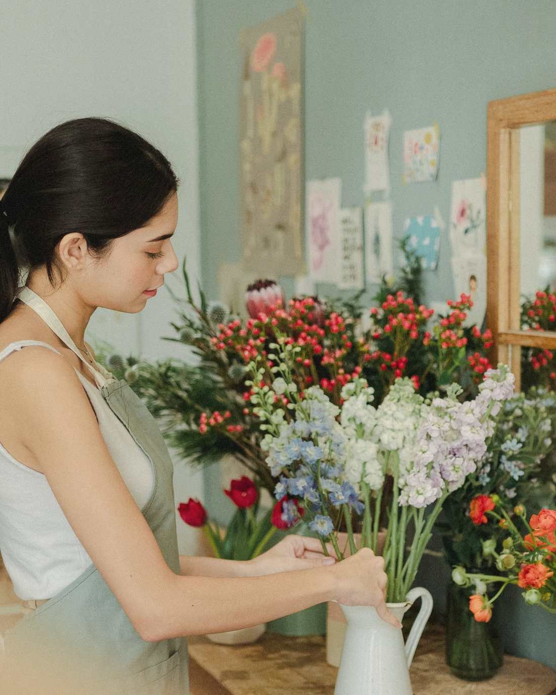 A florist in an apron arranging stocks and roses into a white enamel jug at a wooden counter, surrounded by buckets of flowers.