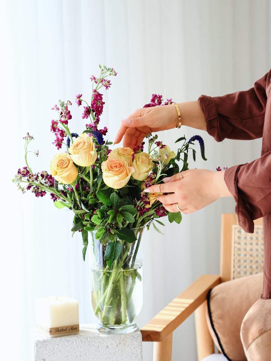 Hands settling yellow roses, purple stocks and greenery into a tall glass vase beside a bright window.