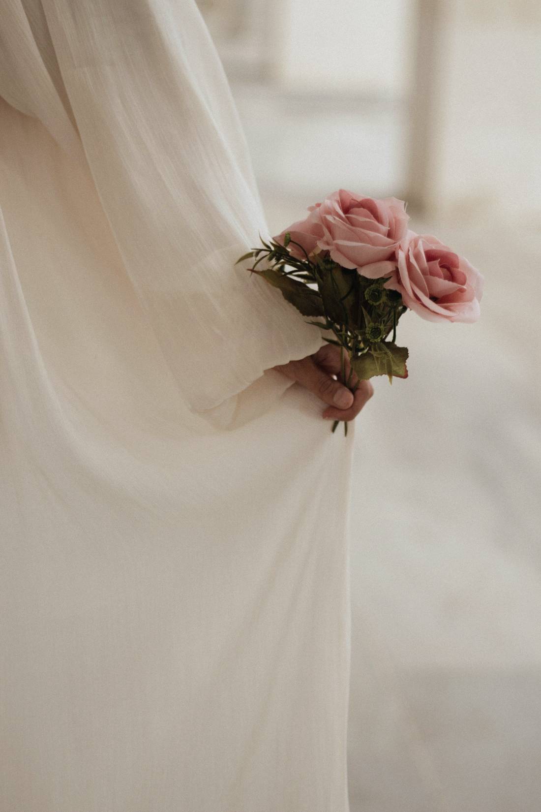 A bride in a flowing ivory gown holding a small, loose posy of dusty pink roses at her side.