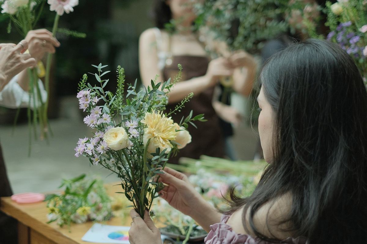 People around a long worktable building small hand-tied bouquets of roses, asters and eucalyptus.